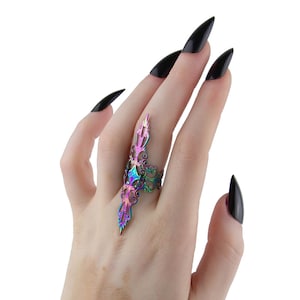 Puede incluir: Un anillo de dedo iridiscente único con un diseño decorativo alargado. El anillo presenta una mezcla de tonos morados, rosas y verdes, creando un efecto visual llamativo. El diseño del anillo se extiende a lo largo del dedo, añadiendo un toque audaz.