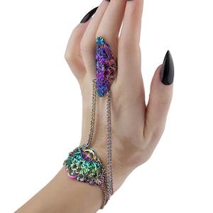 May include: A hand wearing a unique piece of jewelry. The ring and bracelet are connected by a chain. The ring features a large, iridescent stone with purple, blue, and green hues. The bracelet has a matching design. The nails are painted black.