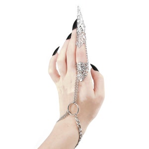 Pu&ograve; includere: Un anello a forma di artiglio in argento con una catena che si estende fino al polso. L'anello &egrave; indossato sull'indice e la catena &egrave; attaccata a un piccolo anello in argento al polso.