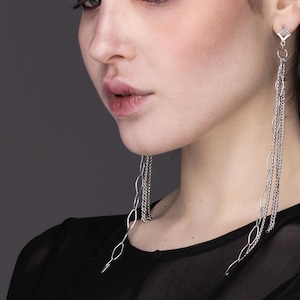 May include: A woman with dark hair and red eyeshadow wears long silver chain earrings. The earrings are made of multiple chains of different sizes and styles.