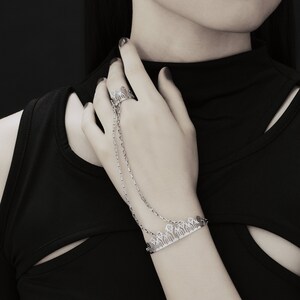 May include: A silver chain bracelet with a delicate, intricate design. The bracelet is worn on the wrist of a person wearing a black top.