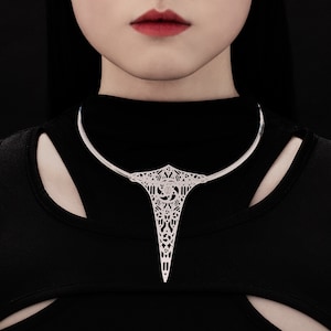 May include: A silver statement necklace with a geometric design. The necklace is made of a thin metal band that circles the neck and has a large, intricate pendant that hangs down the front. The pendant is shaped like a teardrop and has a delicate, filigree design.