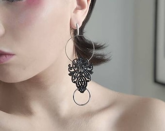 May include: A woman with short black hair wearing black dangle earrings with intricate designs. The earrings feature multiple circles and a delicate, lacy pattern.