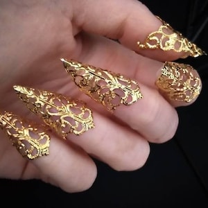May include: Five gold-colored claw rings with intricate filigree designs. The rings are worn on the fingers of a hand.