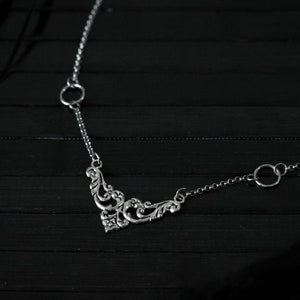 May include: A silver necklace with a delicate, ornate pendant and two circular links. The necklace is on a black wooden surface.