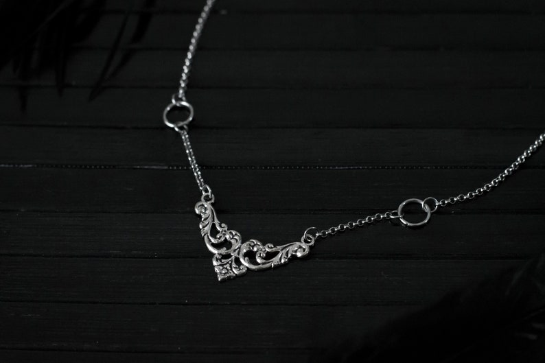 May include: A silver necklace with a delicate, ornate pendant and two circular links. The necklace is on a black wooden surface.