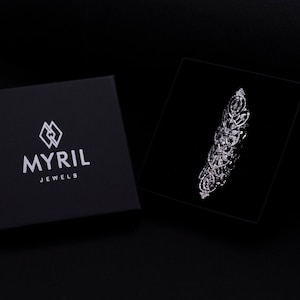 Op de afbeelding: Een zwarte juwelendoos met het logo "MYRIL JEWELS" in wit gedrukt. De doos is open en een zilveren ring met een gedetailleerd ontwerp is zichtbaar in de doos.