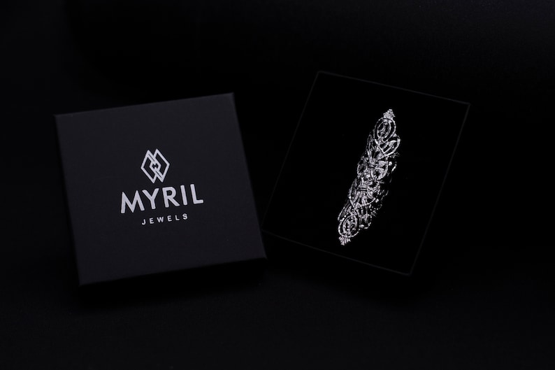 Op de afbeelding: Een zwarte juwelendoos met het logo "MYRIL JEWELS" in wit gedrukt. De doos is open en een zilveren ring met een gedetailleerd ontwerp is zichtbaar in de doos.