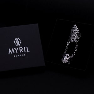 May include: A silver ring with a detailed design and a chain connecting the top and bottom of the ring. The ring is displayed on a black velvet background in a box with the logo "MYRIL JEWELS".