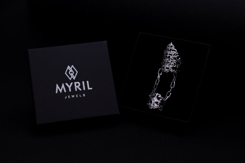 May include: A silver ring with a detailed design and a chain connecting the top and bottom of the ring. The ring is displayed on a black velvet background in a box with the logo "MYRIL JEWELS".