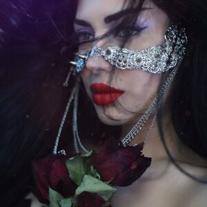 Puede incluir: Una mujer con el pelo largo y negro lleva un aro de nariz de cadena plateada con un dise&ntilde;o decorativo. Est&aacute; sosteniendo un ramo de rosas rojo oscuro.