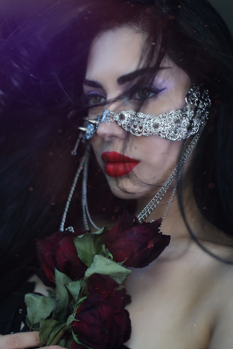 Puede incluir: Una mujer con el pelo largo y negro lleva un aro de nariz de cadena plateada con un dise&ntilde;o decorativo. Est&aacute; sosteniendo un ramo de rosas rojo oscuro.