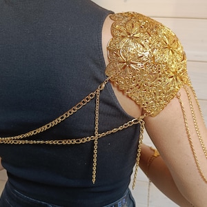 May include: Gold chain shoulder piece with a decorative floral design. The chain is connected to a smaller chain that wraps around the chest.