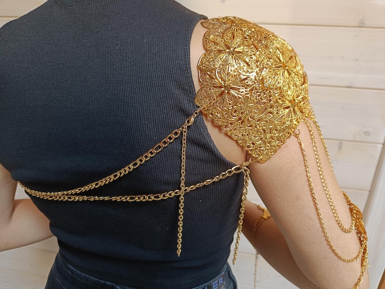 May include: Gold chain shoulder piece with a decorative floral design. The chain is connected to a smaller chain that wraps around the chest.