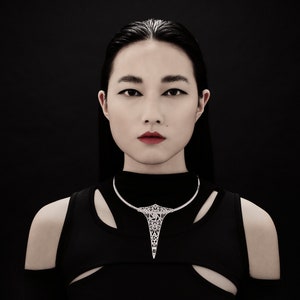 May include: A woman with dark hair and a black top is wearing a silver geometric necklace. The necklace has a sharp point at the bottom.