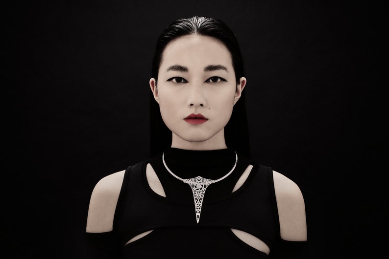 May include: A woman with dark hair and a black top is wearing a silver geometric necklace. The necklace has a sharp point at the bottom.