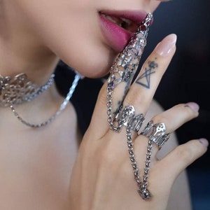 Könnte beinhalten: Silberfarbene, bewegliche Fingerrüstung und passendes Choker-Halsband. Die Fingerrüstung weist aufwendige Ausschnitte auf und ist durch eine Kette verbunden. Der Schmuck wird vor dunklem Hintergrund präsentiert.