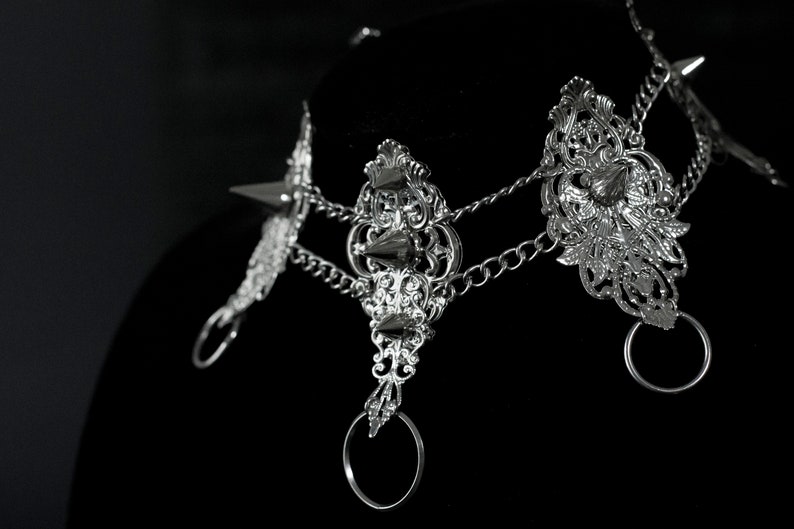 May include: Silver choker necklace with ornate details, featuring a chain link design and multiple spikes. The necklace has three decorative rings.