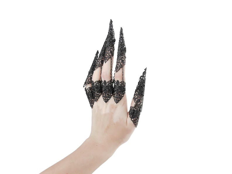 May include: A hand wearing a set of black, intricate, metal finger rings. The rings are designed to resemble claws or talons, with delicate, lacy details.