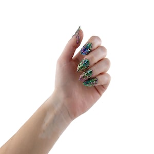 Puede incluir: Una mano con cinco anillos de u&ntilde;as iridiscentes con un dise&ntilde;o floral. Los anillos son puntiagudos y tienen un acabado met&aacute;lico.