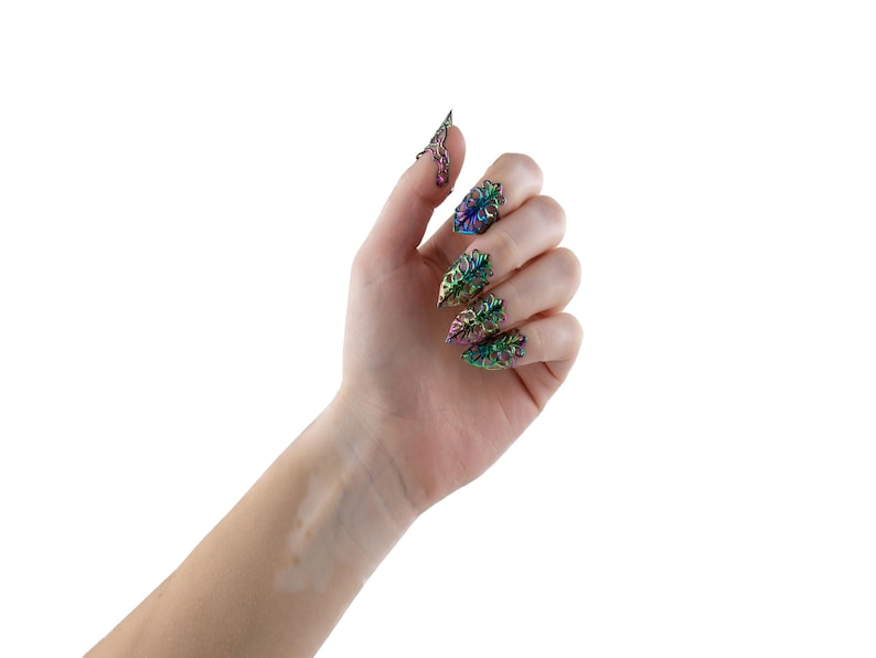 Puede incluir: Una mano con cinco anillos de u&ntilde;as iridiscentes con un dise&ntilde;o floral. Los anillos son puntiagudos y tienen un acabado met&aacute;lico.
