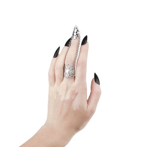 Puede incluir: Un anillo de plata con un delicado dise&ntilde;o floral y una cadena que se extiende hasta el dedo de al lado. El anillo se lleva en una mano con esmalte de u&ntilde;as negro.