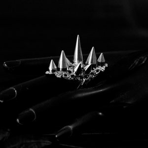 Pu&ograve; includere: Un anello d'argento con pi&ugrave; punte appuntite, che ricorda una corona, su uno sfondo nero.