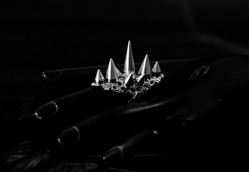 Pu&ograve; includere: Un anello d'argento con pi&ugrave; punte appuntite, che ricorda una corona, su uno sfondo nero.