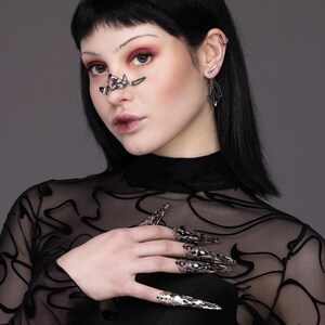 May include: A person wearing a black mesh top with silver claw-like rings on their fingers. The rings are detailed and intricate.