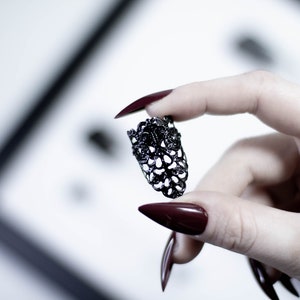 May include: A black metal ring with a delicate, intricate design. The ring is held between two fingers with dark red nail polish.