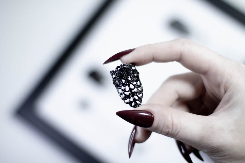 May include: A black metal ring with a delicate, intricate design. The ring is held between two fingers with dark red nail polish.