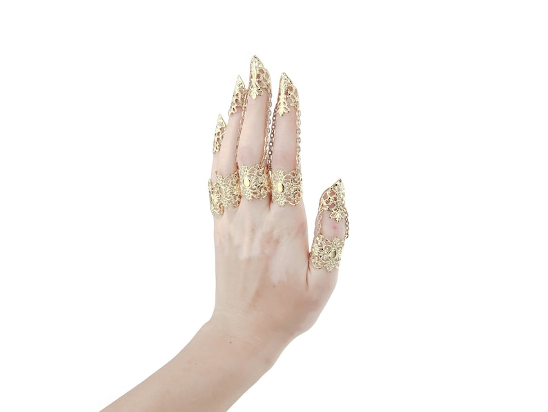 Puede incluir: Un conjunto de cinco anillos de dedo de tono dorado con intrincados dise&ntilde;os de filigrana. Los anillos est&aacute;n conectados por cadenas y presentan puntas puntiagudas.