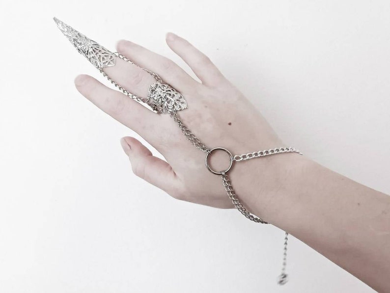 Pu&ograve; includere: Un anello d'argento con un design a punta e filigrana e una catena che si collega a un braccialetto d'argento con un ciondolo circolare.