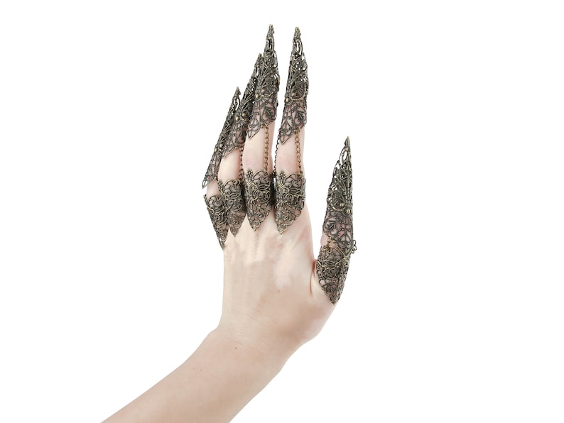 May include: A hand wearing five ornate, gold-toned finger rings. The rings are designed to resemble claws and are intricately detailed with a filigree pattern.