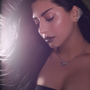 May include: A woman with long black hair wearing a black strapless top and a silver necklace with a floral design. She has dark makeup and a silver nose ring.