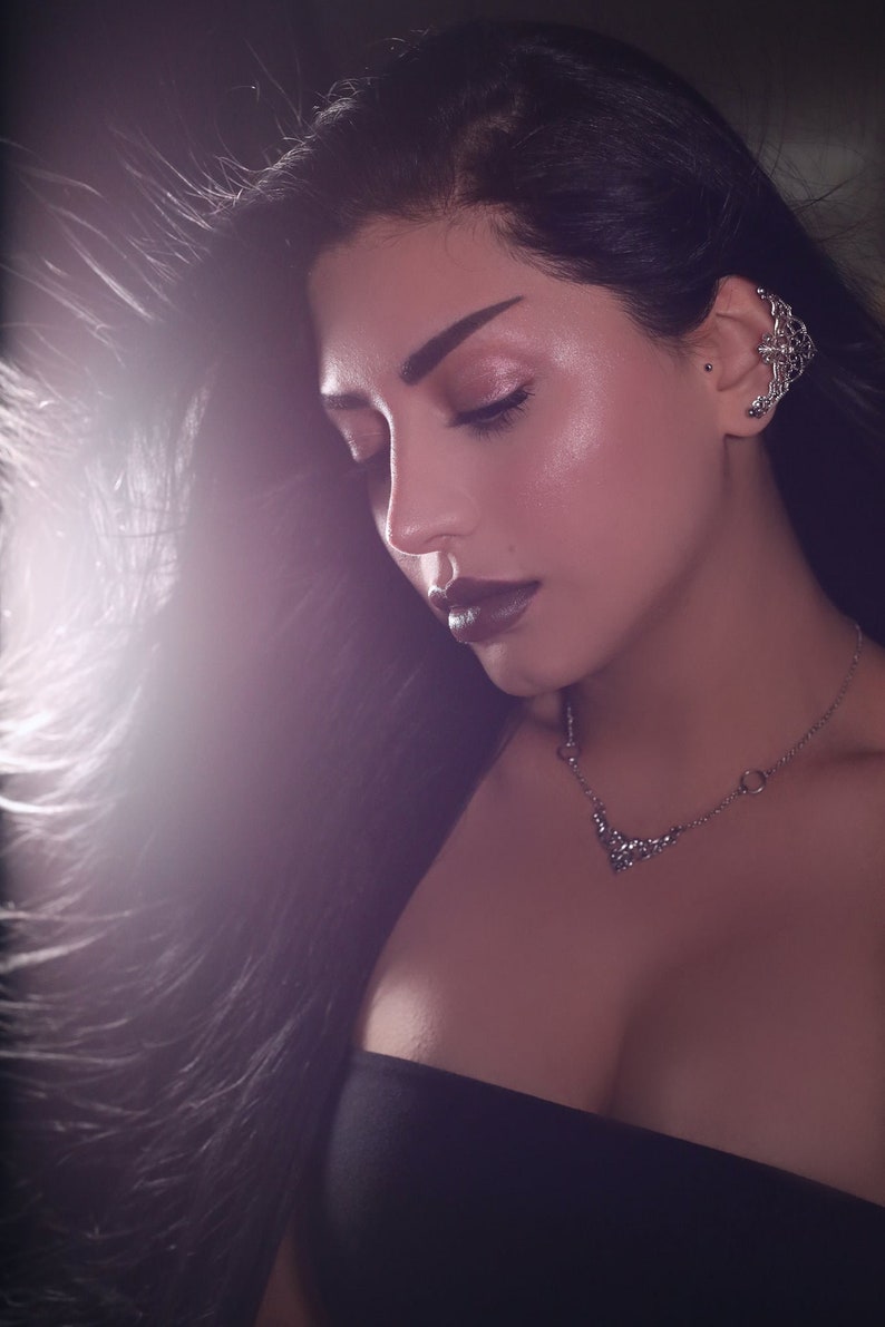 May include: A woman with long black hair wearing a black strapless top and a silver necklace with a floral design. She has dark makeup and a silver nose ring.
