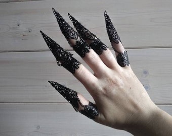 May include: Black lace finger rings with pointed tips, resembling claws. The rings are worn on multiple fingers of a hand.