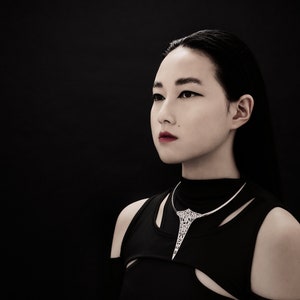 May include: A woman with long black hair wearing a black mock neck shirt and a silver necklace with a geometric design.