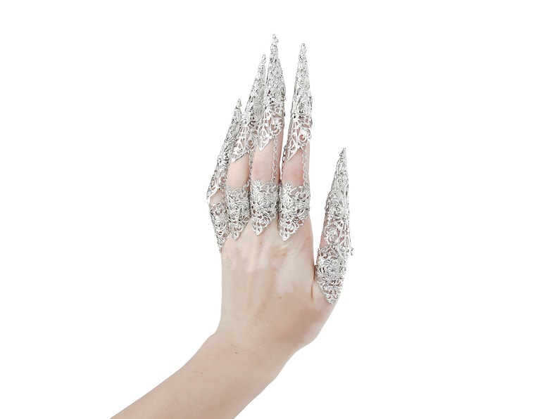May include: A hand wearing silver filigree claw rings. The rings are intricate and detailed, with a pointed design.