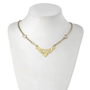 May include: A gold necklace with a delicate, ornate pendant in the shape of a stylized heart. The pendant features intricate floral designs and is connected to a thin gold chain.