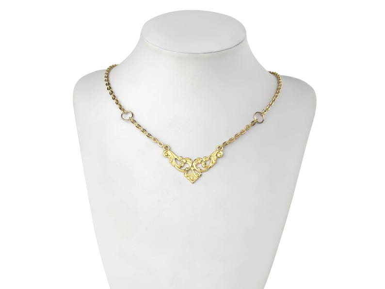 May include: A gold necklace with a delicate, ornate pendant in the shape of a stylized heart. The pendant features intricate floral designs and is connected to a thin gold chain.