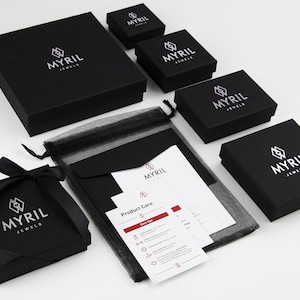 Puede incluir: Cajas de joyer&iacute;a negras con el logotipo "Myril Jewels" impreso en blanco. Las cajas est&aacute;n dispuestas en forma de pir&aacute;mide con una bolsa de malla negra y una tarjeta blanca con instrucciones de cuidado del producto.
