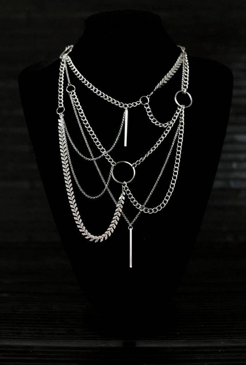 May include: A silver layered necklace with multiple chain styles, including a herringbone chain, and two pendant bars. The necklace features two circular rings.