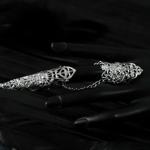 May include: Two silver filigree rings connected by a chain. The rings are designed to be worn on two fingers. The rings are on a black mannequin hand.