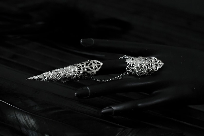 May include: Two silver filigree rings connected by a chain. The rings are designed to be worn on two fingers. The rings are on a black mannequin hand.