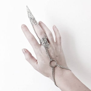Pu&ograve; includere: Un anello d'argento con una lunga protezione per le dita a punta e una catena che si estende fino al polso. La catena ha un anello circolare al polso.