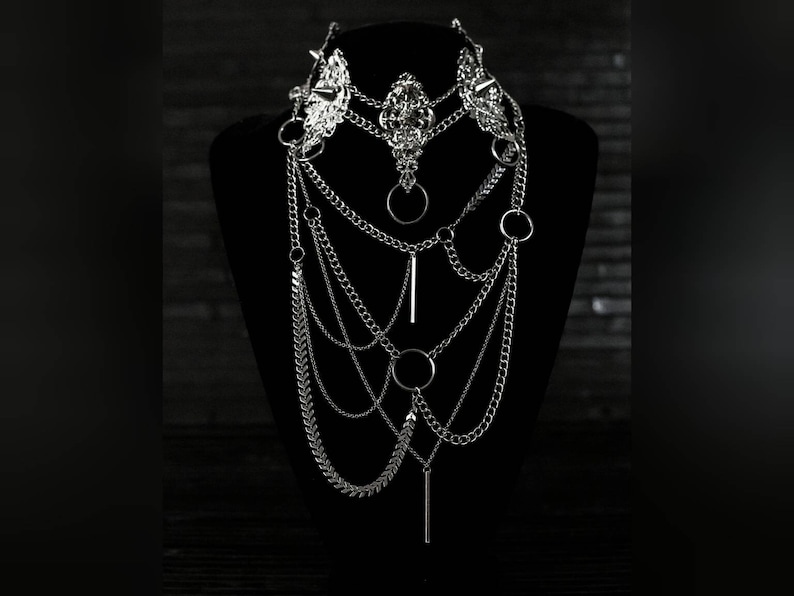 May include: A silver layered chain necklace with multiple chain styles, including a delicate chain, a curb chain, and a chain with small spikes. The necklace features several silver rings and a long silver bar pendant.