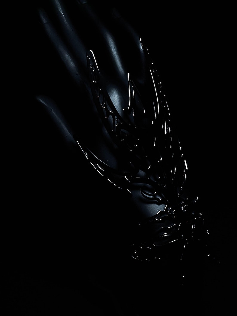Puede incluir: Una escultura de metal negro con detalles intrincados, que se asemeja a una mano esquel&eacute;tica o un p&aacute;jaro estilizado en vuelo. La escultura est&aacute; iluminada desde un lado, proyectando sombras dram&aacute;ticas.