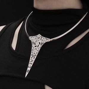 May include: A silver choker necklace with a delicate, intricate design. The necklace features a long, pointed pendant that hangs down the front of the neck.