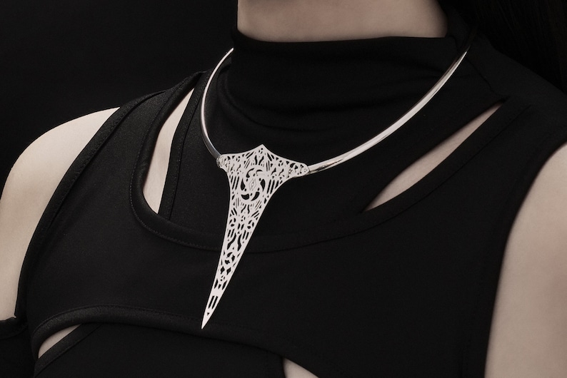 May include: A silver choker necklace with a delicate, intricate design. The necklace features a long, pointed pendant that hangs down the front of the neck.
